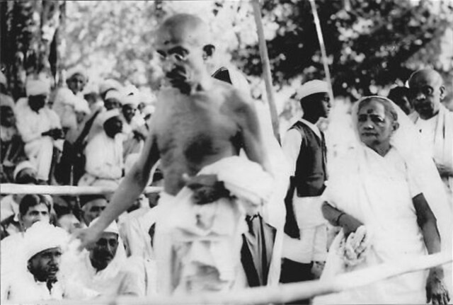 A black and white image of Mahatma Gandhi in front of a crowd. He seems to be walking past the crowd.