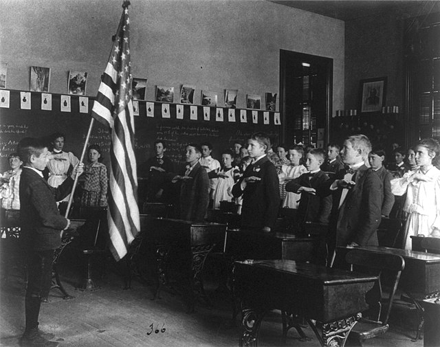 Black and white image of students in formal uniforms standing and taking the Pledge of Allegiance. One student in the far left of the photo is holding the flag of the United States at the the front of classroom. The rest of the students and teacher are facing him with their hands over their hearts.