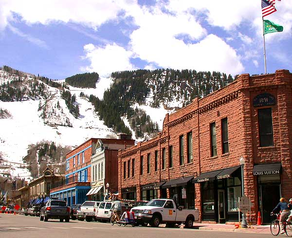 An image of downtown Aspen, Colorado, with brick buildings housing stores against the backdrop of a mountain that has snow and trees. Cars and trucks are parked in front of the stores.