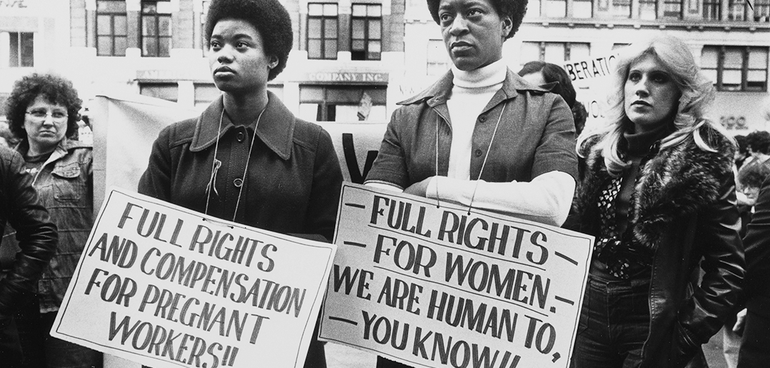 The center of the image depicts two black women holding protest signs that read "full rights and compensation for pregnant workers!!" and "full rights for women. We are human to, you know. White women behind the center protesters are holding a banner. Other signs saying "liberation" can be seen in the background.