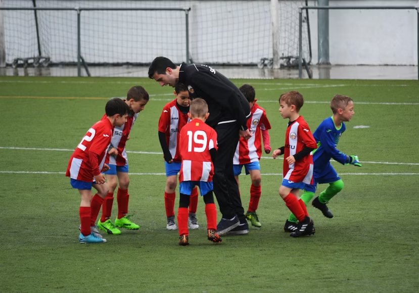 An image of young boys in red, white, and blue soccer uniforms in a circle around their coach, a man dressed in black. One of the children dressed in blue goalie gear is running off.