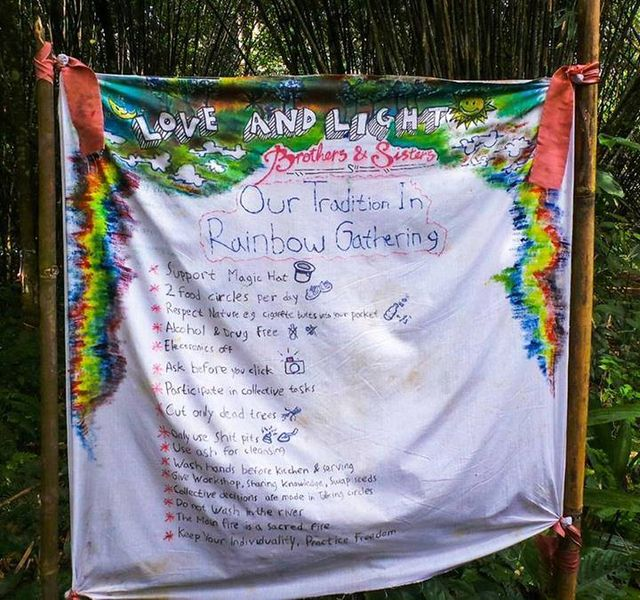 Image of a sheet tied to two polls. The top and half way down the sides is tie dyed in rainbow colors. In the tie dye at the top it reads Love and Light, Brothers and Sisters. Then in larger text "Our Tradition in the Rainbow Gathering" underneath that in smaller text it reads: support magic hat, 2 food circles per day, respect nature, alcohol and drug free, electronics off, ask before you click, participate in collective tasks, cut only dead trees, only use shit pits, use ash for cleansing, wash hands before kitchen and serving, give workshop sharing knowledge swap seeds, collective decisions are made in talking circles, do not wash in the river, the main fire is a sacred fire, keep your individuality practice freedom