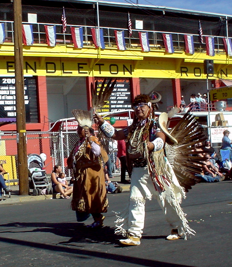 Man with dark skin tone in tribal regalia walks in a parade. Families sit and stand observing the parade.