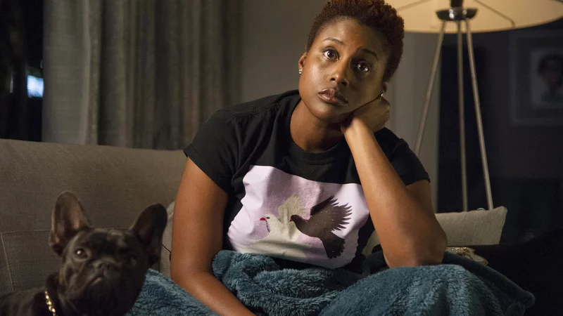 Image of Issa Rae, a person with a dark skin tone and short hair, sitting on a couch with one hand on her shoulder, next to a small dog.