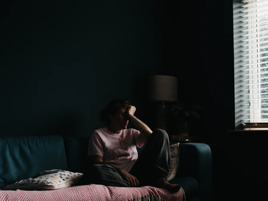 A person in a pink shirt and grey pants sitting on a couch in a dark room, with light streaming through the blinds on the window to the right.