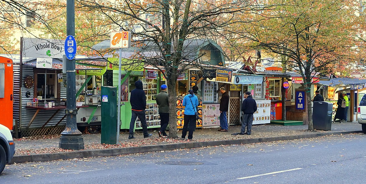 An image of a Portland food cart pod in the fall. Several people wait on the sidewalk in front of the carts.