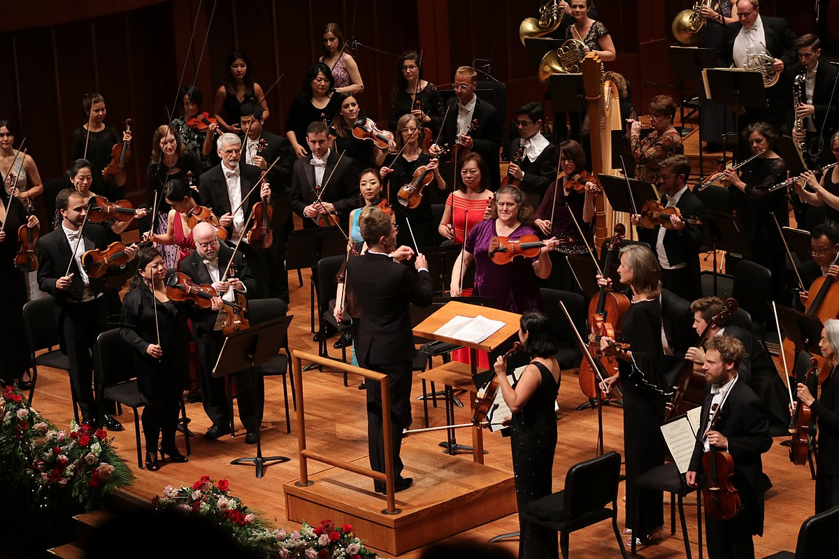 Photo depicting the Indianapolis Symphony Orchestra in formal dress on stage during an opening night gala. The conductor is in the middle of the photo holding a baton, and the musicians have their instruments in their hands.