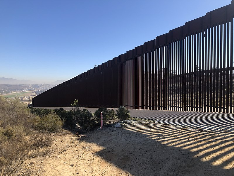 An image of a border wall made of metal slats with the wall casting a shadow onto desert soil. The photo shows a desert landscape and sage brush. In the far distance is an urban area bisected by the wall.