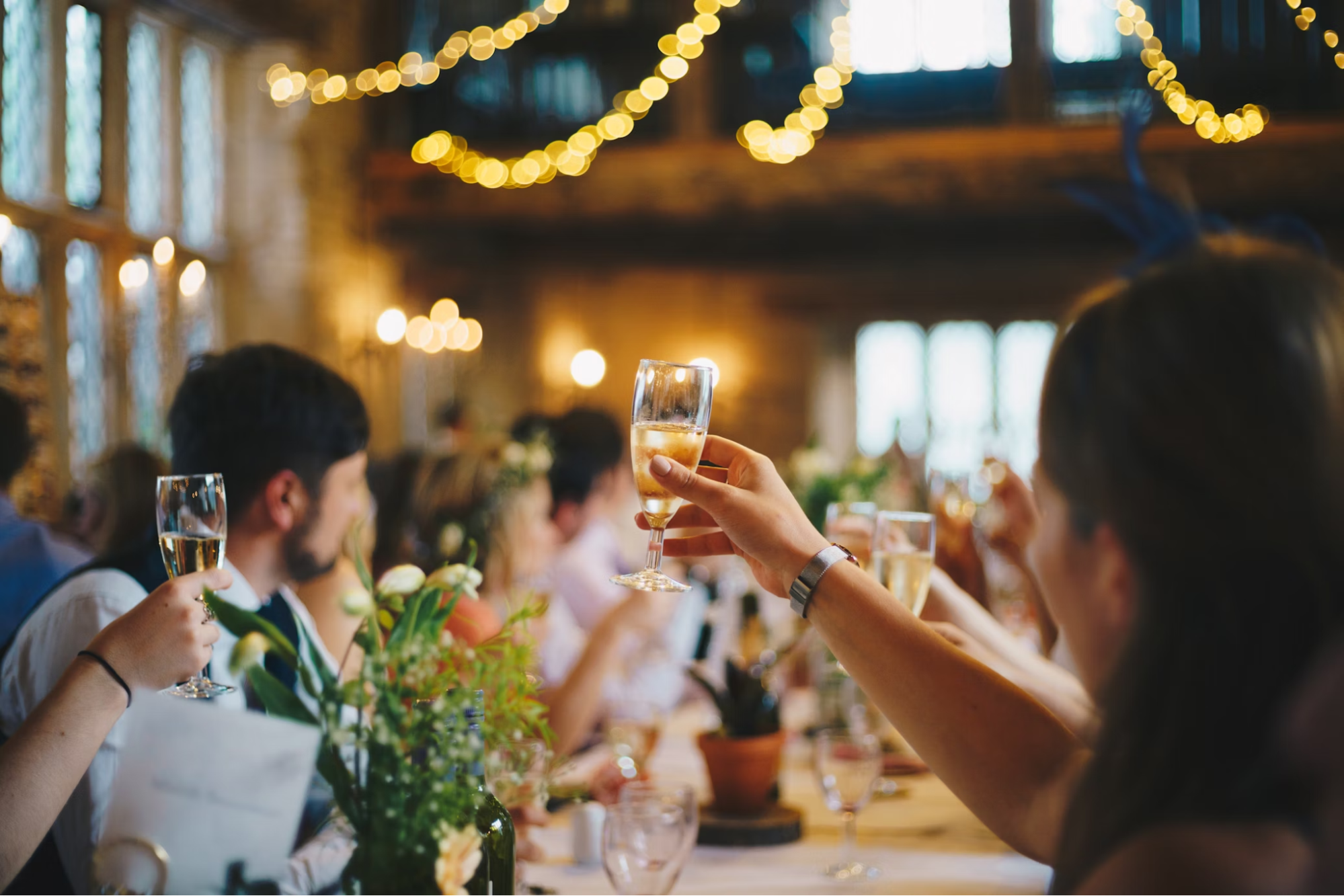 People raising wine glasses at a wedding while sitting at a table that has other glasses and flowers on it, string lights blurred in the background.