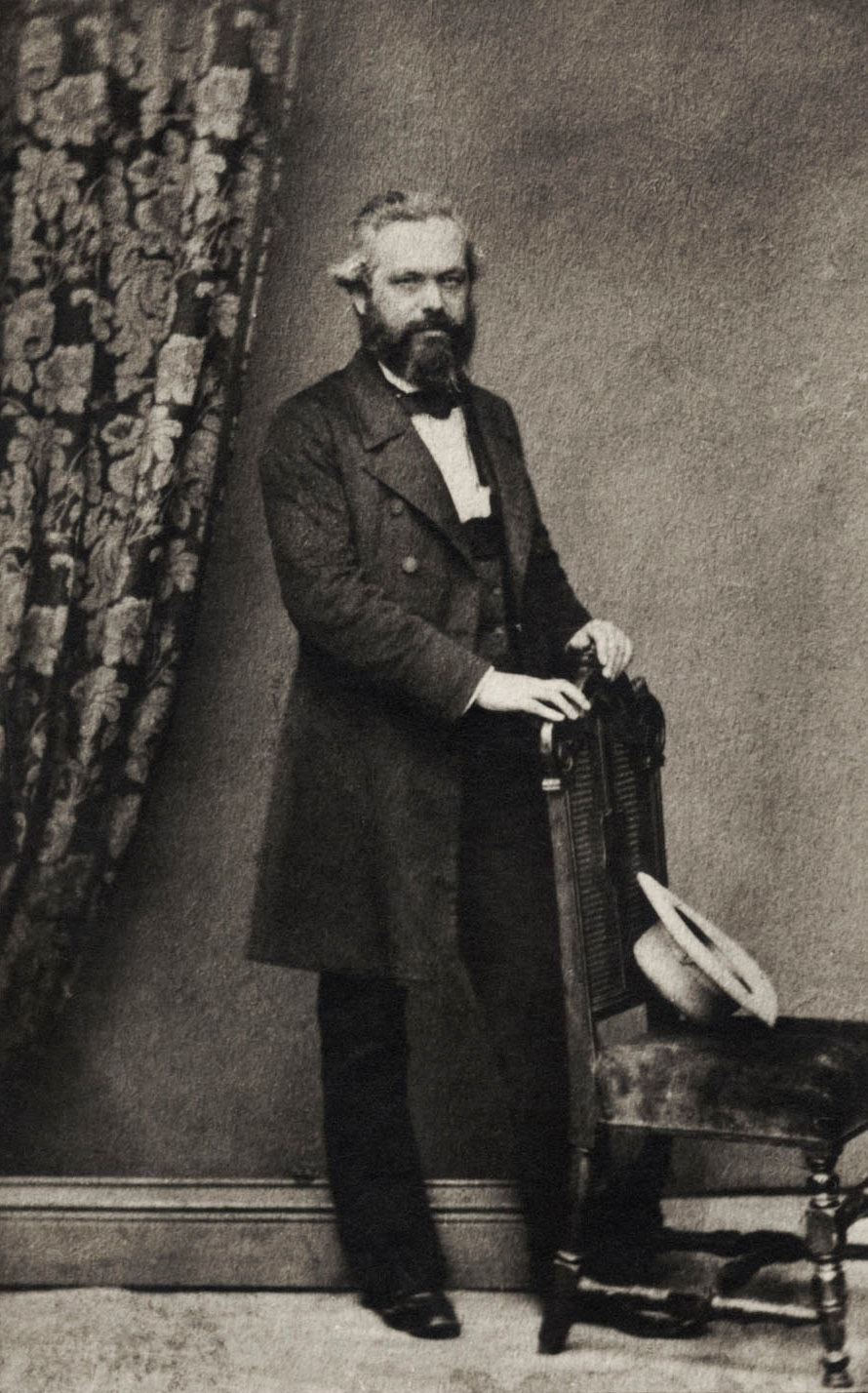 A black and white photograph of Karl Marx in a suit standing behind a chair.