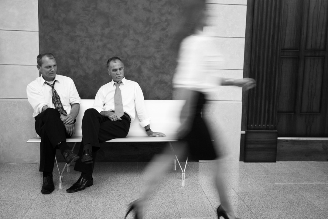 An image of a blurred woman walking in the foreground while being watched by men sitting nearby.