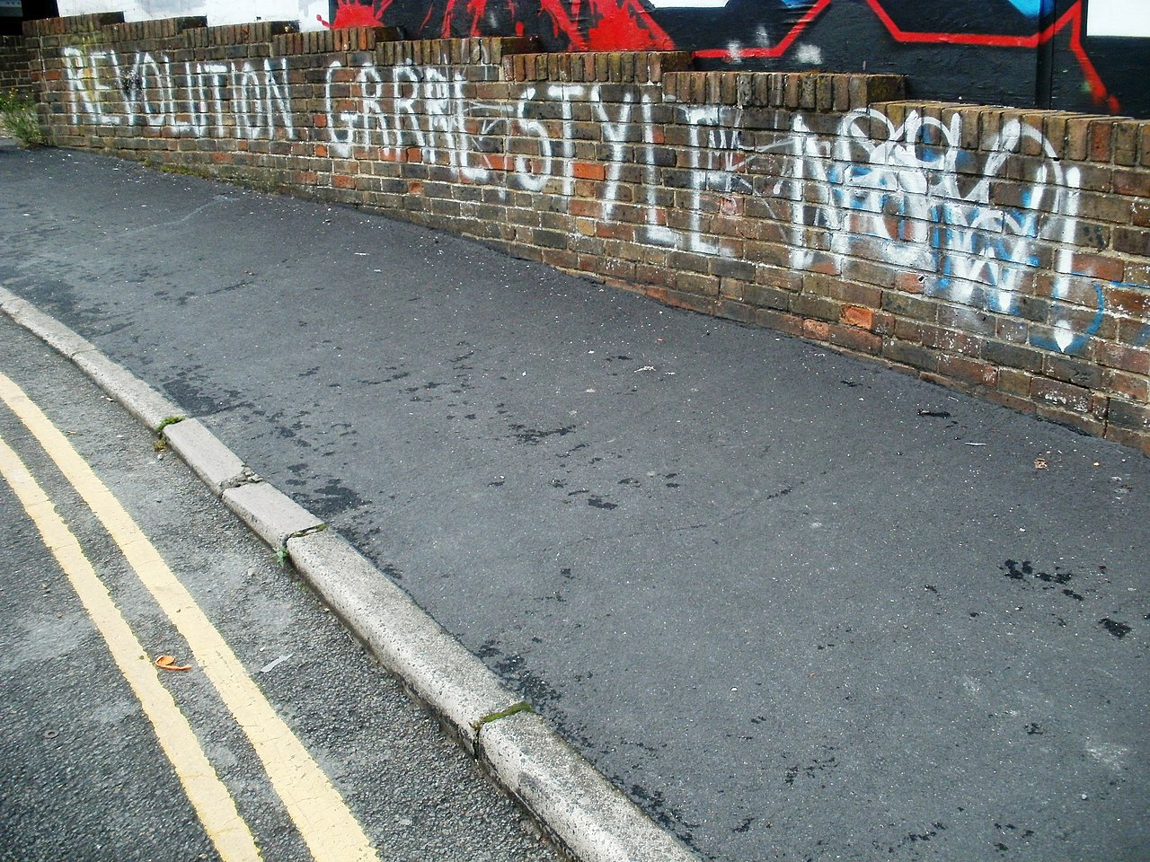 Image of graffiti saying “revolution grrrl style now!” on a brick wall near a road.