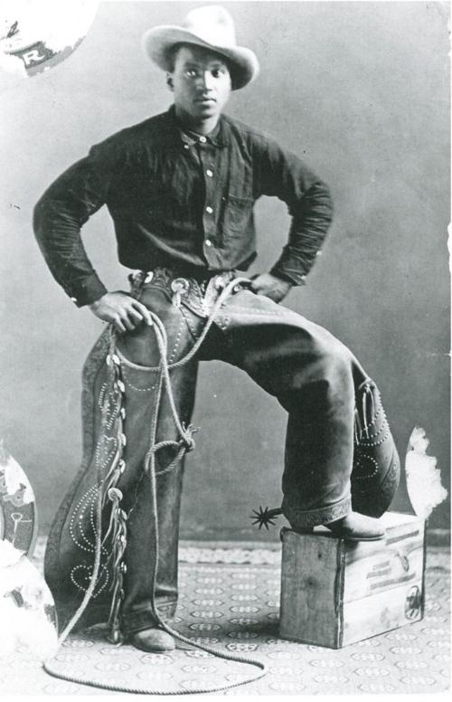 A man with dark skin tone wearing riding chaps and boots with spurs.