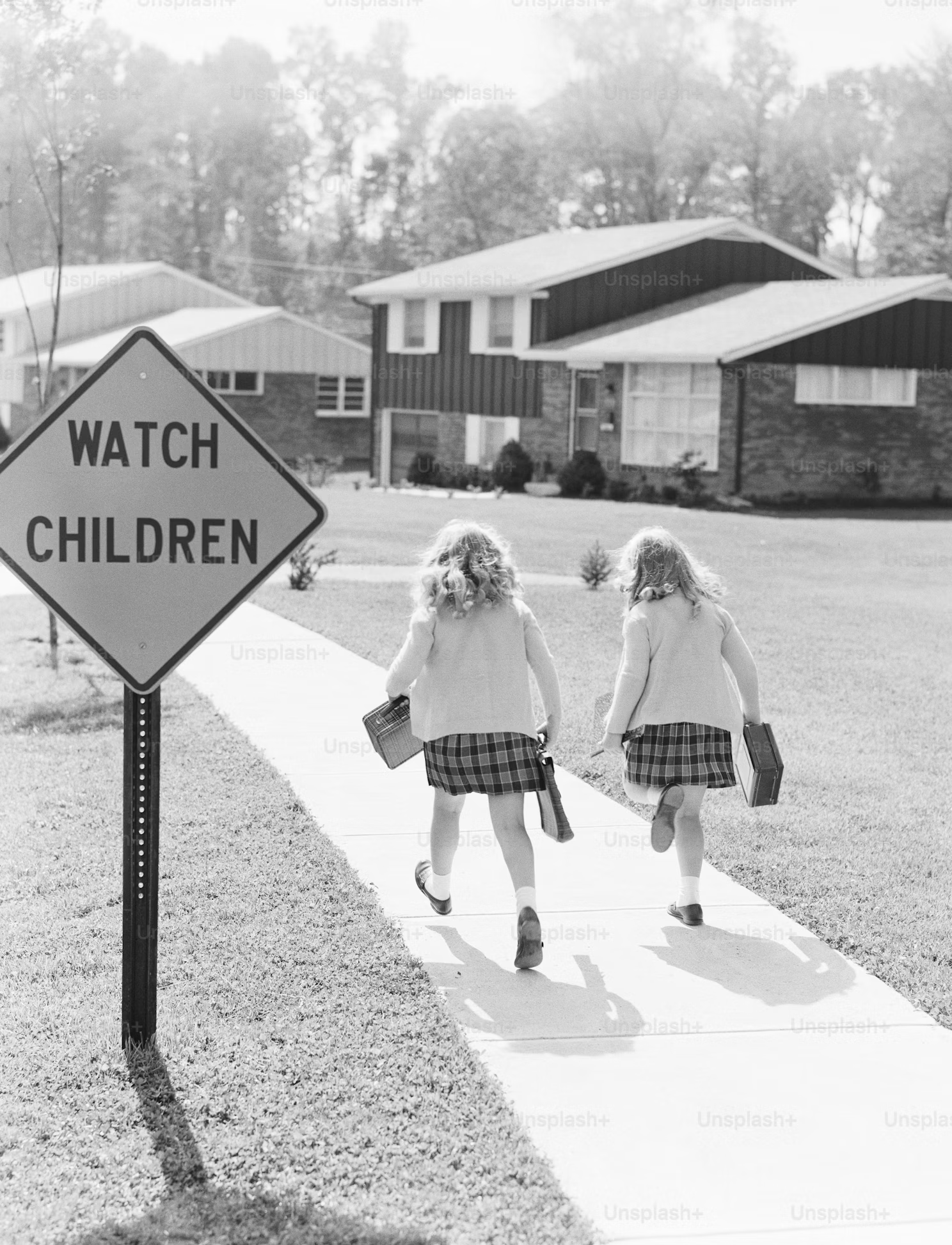 Suburban neighborhood with two young girls walking with lunch boxes or small bags. A sign next to the path says "Watch Children."