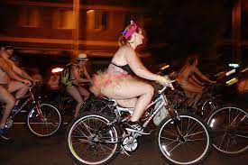 Foreground: A woman with light tone skin riding a bicycle while wearing a triangle top and tutu. Background: Other cyclists with light skin tones in limited clothing biking during an evening protest.