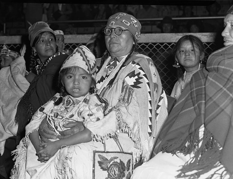 A black and white photo of a tribal elder (woman) holding a young child. Other women and children are seated nearby.