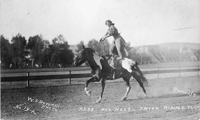 A black and white photo of two women performing a riding trick on a horse.