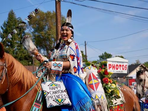 A woman with medium skin tone rides a horse and has a bag that reads "Happy Canyon Princess."