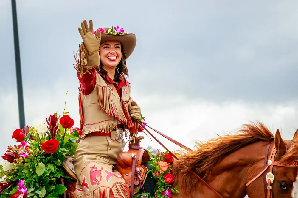 A woman with a light skin tone rides a horse while wearing a western hat with flowers and a fringe vest and skirt.