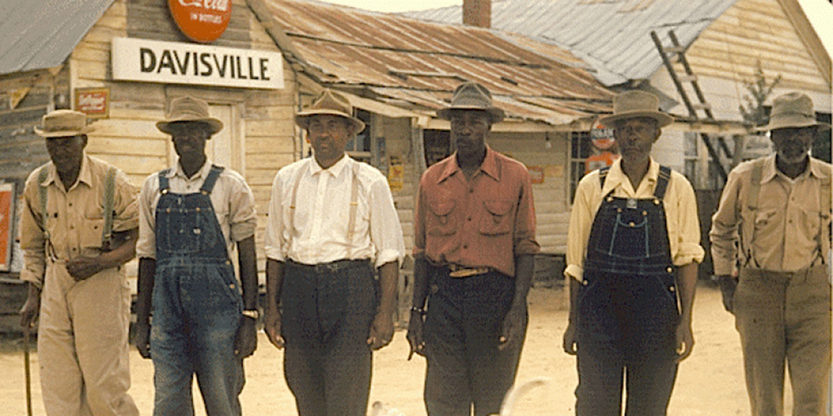 Six men with dark skin tones, some wearing pants with suspenders or overalls, all wearing hats, walk down a dirt street in front of a sign for "Davisville."