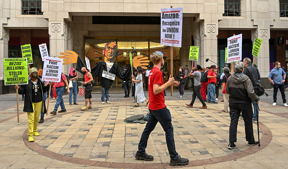 Photo of people marching in a circle around a stone plaza in front of a building. People are from different backgrounds. At the back of the group there is someone dressed as a Jeff Bezos puppet with large hands. The strikers hold signs stating either "Bezos' Billions: Stolen from Workers" on a white board, or "Fight Racism and Union Busting" on neon yellow boards.