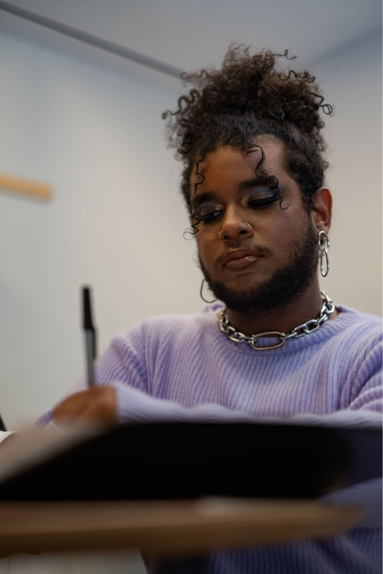 Non-binary person with dark skin tone sitting at a desk writing. Curly hair is in an updo, full face makeup, facial hair (mustache and beard), wearing earrings, a bold necklace, and a lavender sweater