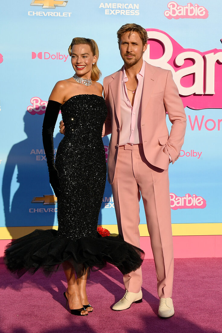 Image of Margot Robbie and Ryan Gosling on a pink carpet in front of a blue wall covered in pink Barbie logos and the logos of other businesses.