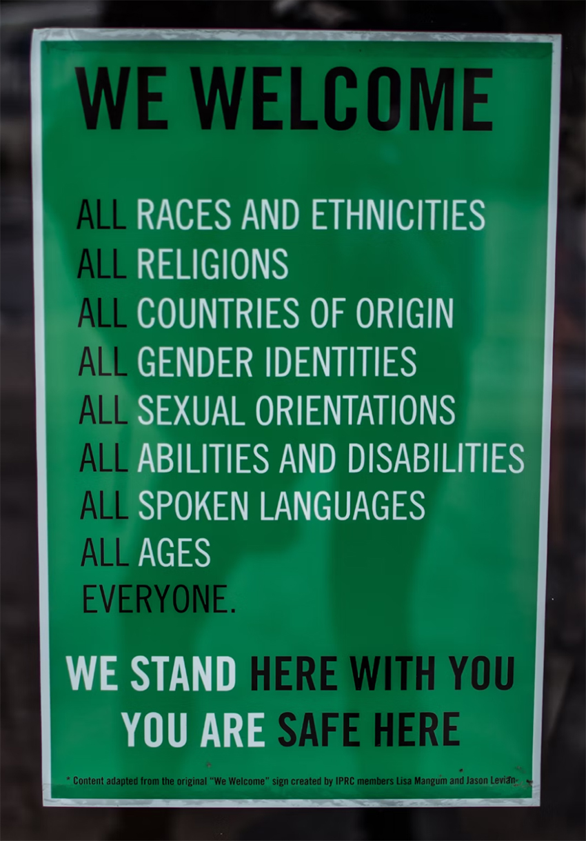 Green sign says "We welcome, all races and ethnicities, all religions, all countries of origin, all gender identities, all sexual orientations, all abilities and disabilities, all spoken languages, all ages, everyone. We stand here with you and you are safe here."