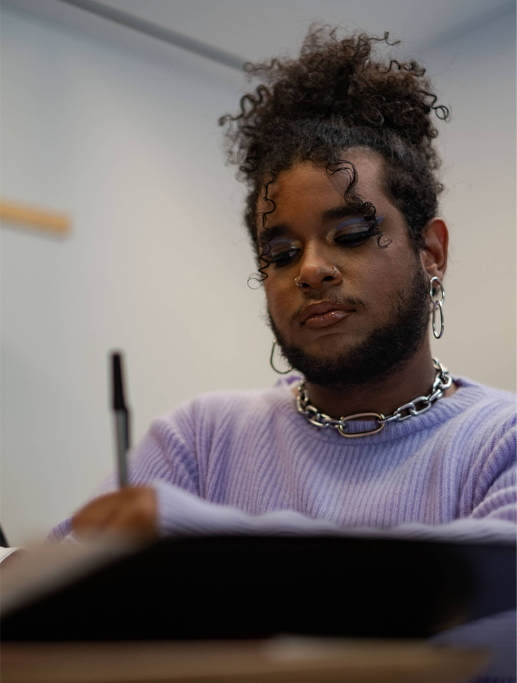 Non-binary person with dark skin tone sitting at a desk writing. Curly hair is in an updo, full face makeup, facial hair (mustache and beard), wearing earrings, a bold necklace, and a lavender sweater