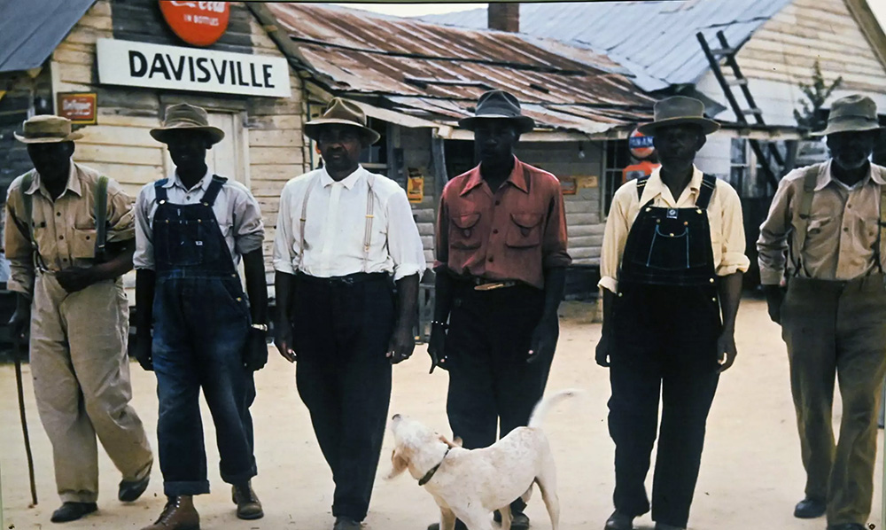 Six men with dark skin tones, some wearing pants with suspenders or overalls, all wearing hats, walk down a dirt street in front of a sign for "Davisville."