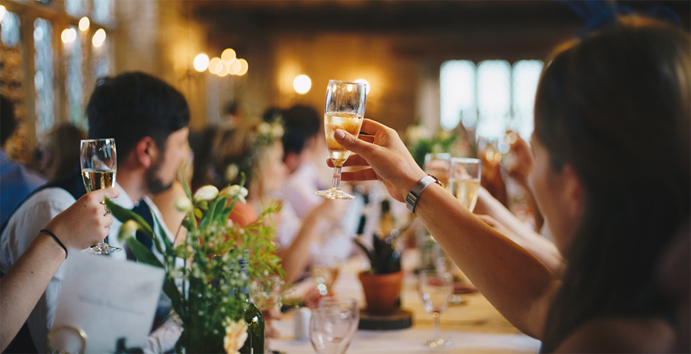 People raising wine glasses at a wedding while sitting at a table that has other glasses and flowers on it, string lights blurred in the background.