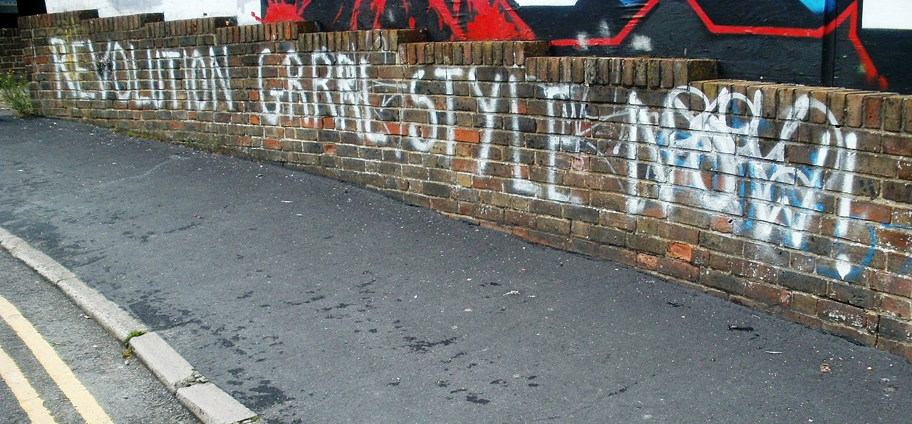 Image of graffiti saying “revolution grrrl style now!” on a brick wall near a road.