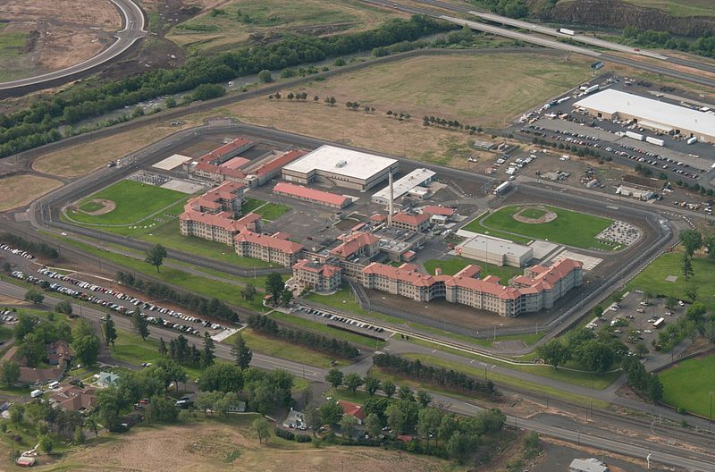 An aerial view of Eastern Oregon Correctional Institution in Pendleton, Oregon. A large, multi-winged building structure with a red roof and tan brick walls. Areas of green grass are fenced off. Situated in a rural location with roads nearby.
