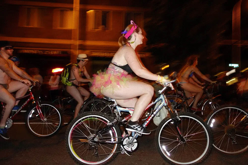 Foreground: A woman with light tone skin riding a bicycle while wearing a triangle top and tutu. Background: Other cyclists with light skin tones in limited clothing biking during an evening protest.