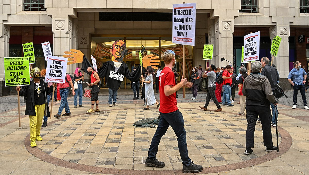 Photo of people marching in a circle around a stone plaza in front of a building. People are from different backgrounds. At the back of the group there is someone dressed as a Jeff Bezos puppet with large hands. The strikers hold signs stating either "Bezos' Billions: Stolen from Workers" on a white board, or "Fight Racism and Union Busting" on neon yellow boards.