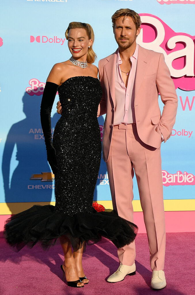 Image of Margot Robbie and Ryan Gosling on a pink carpet in front of a blue wall covered in pink Barbie logos and the logos of other businesses.