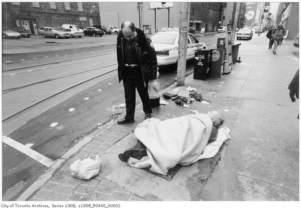 A police officer stands over a person who is trying to sleep on the street, covered by a blanket.