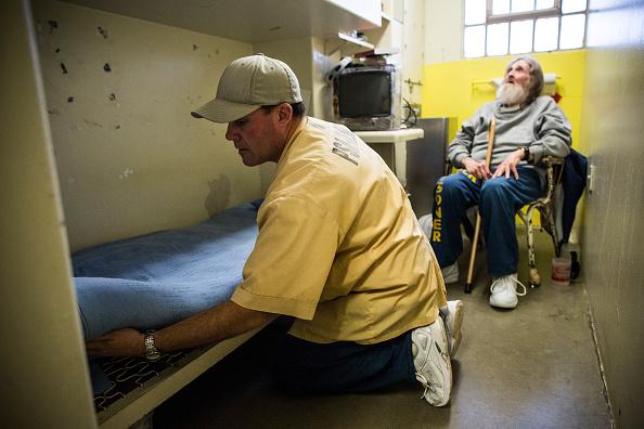 A inmate makes the bed of an older inmate sitting in a chair with a cane.