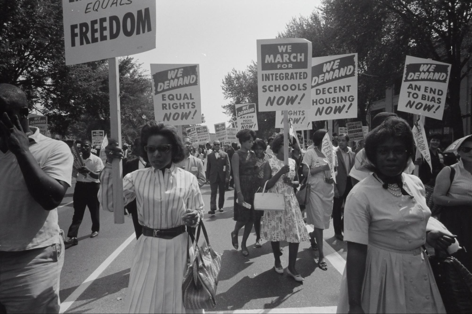 Photo of Black protesters holding signs: "We demand equal rights now!" and "We march for integrated schools now!" and "We demand decent housing now!" and "We demand and end to bias now!"