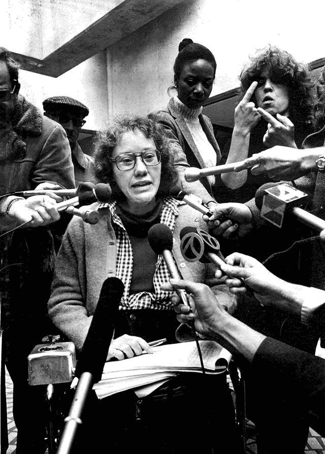 Photo of Kitty Cone surrounded by microphones held by reporters at a press conference