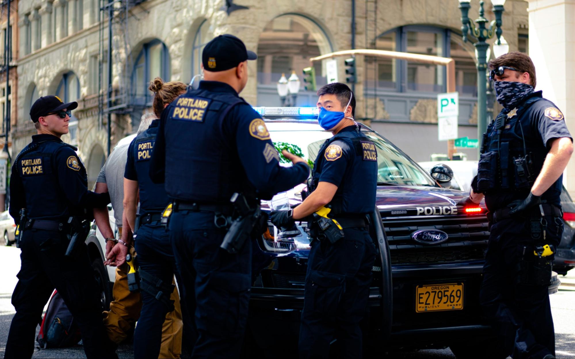 Police officers in uniform and face masks gather around a policy car. A person in handcuffs is visible behind two officers, and a third officer holds the handcuffed person's arm.