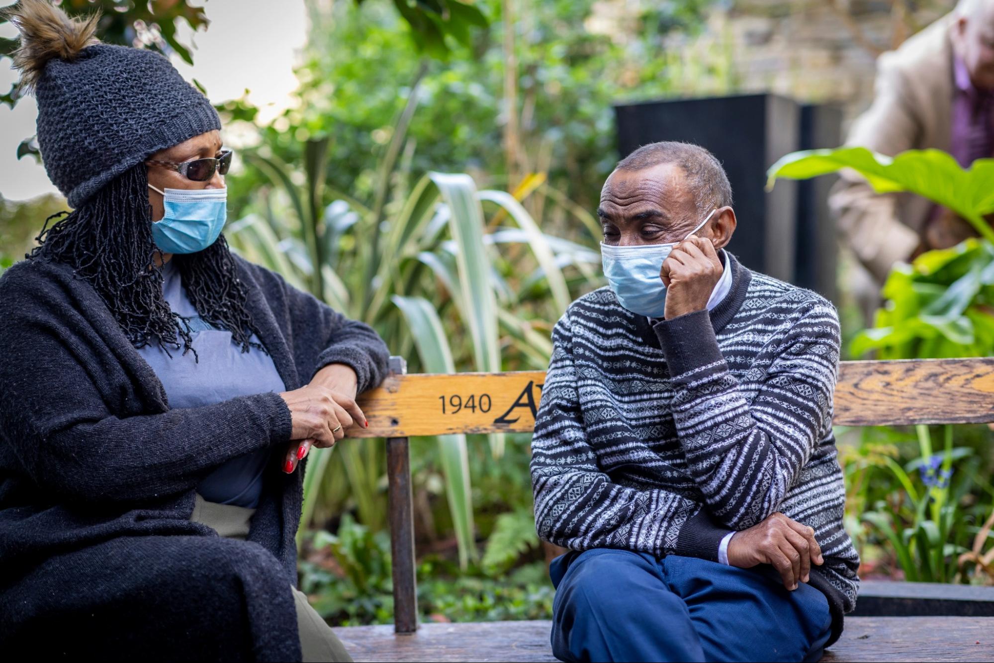 Two people talking on a park bench. Both wear masks.