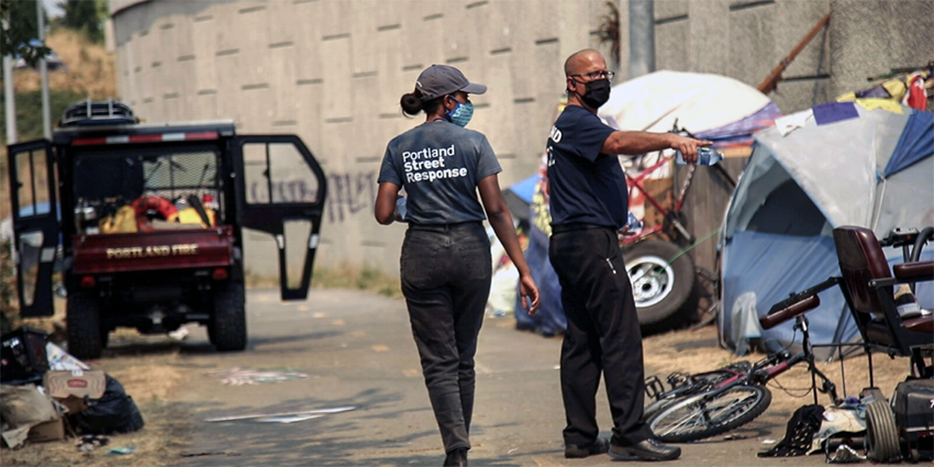 Two Portland Street Response workers wearing face masks in an urban environment with a bicycle and tents.