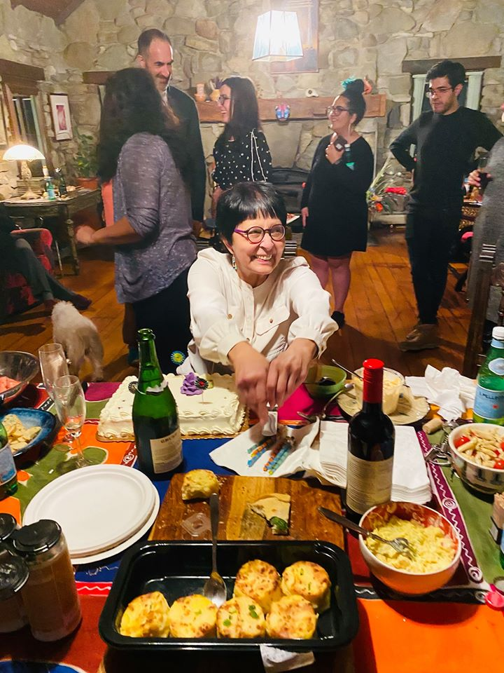 Maria Lugones, An older feminine-presenting person with brown skin and short brown hair sits at a colorful table full of food and wine. She is smiling and reaching for something on the table, as if she is about to serve her guests. In the background, people are greeting one another.