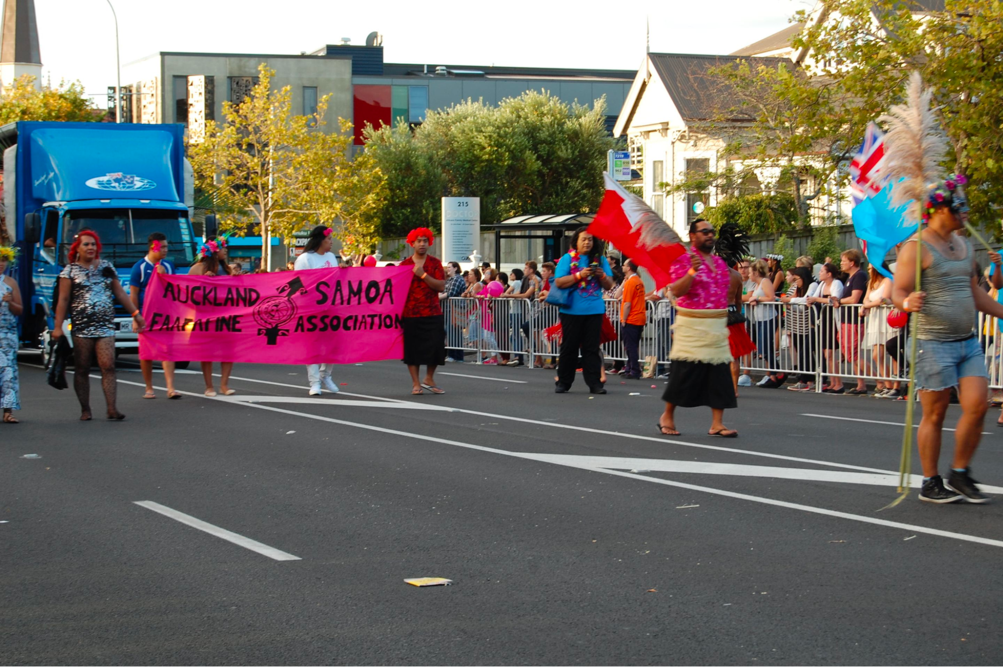 A parade of people in both modern and traditional Samoan clothing carrying a large pink banner with a nonbinary gender symbol, that says, "Aukland, Samoa Fa’afafine Society". Other participants are carrying flags and other symbols of Samoan culture.