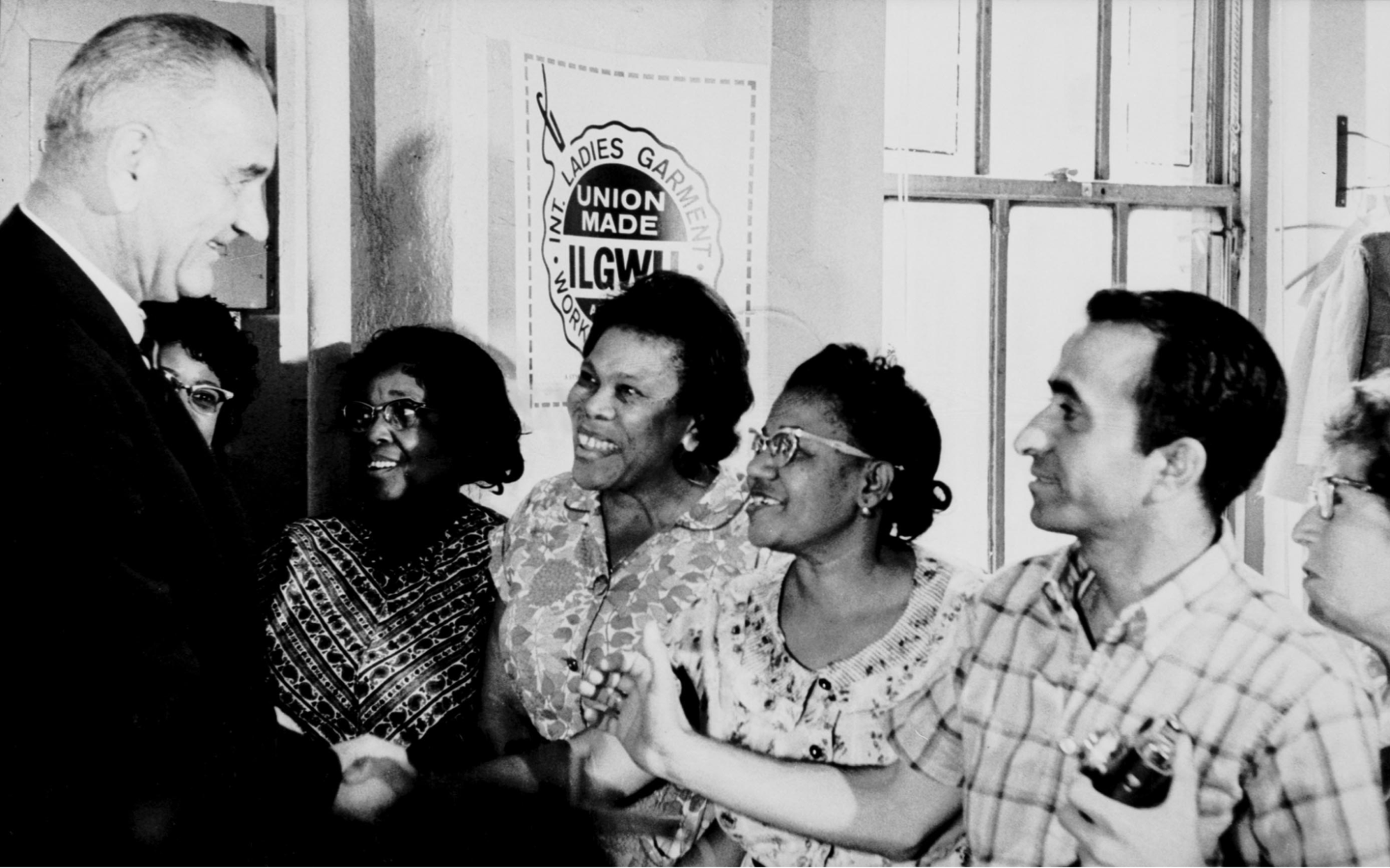 Black and white photo of President Johnson greeting four Black women under a sign for the International Ladies Garment Workers Union. A male photographer is also pictured.