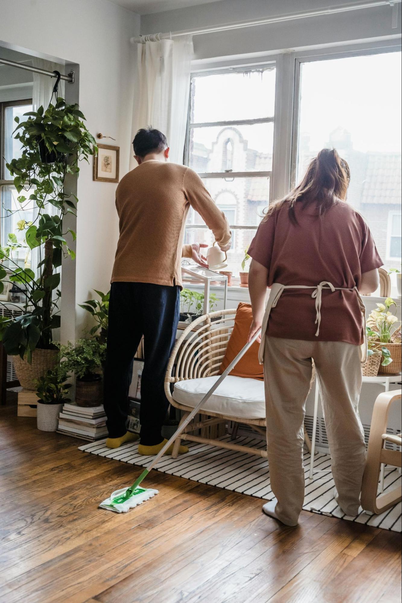 A masculine presenting person and a feminine presenting person cleaning a sunlit room.