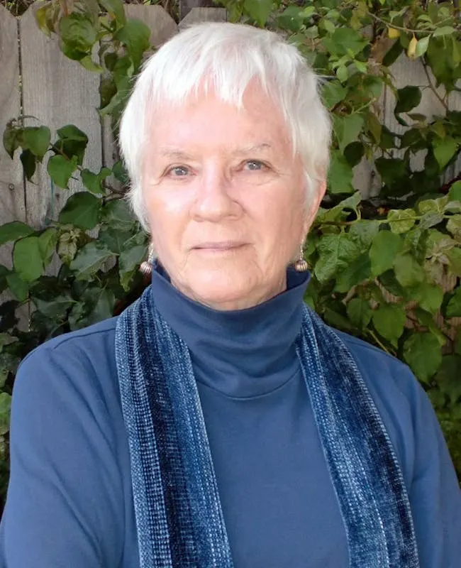 A headshot of Sandra Harding, an older, light-skinned, feminine presenting person with white hair, wearing a blue turtleneck and a blue velour scarf. An old wood fence and green shrubbery suggest she is outside.