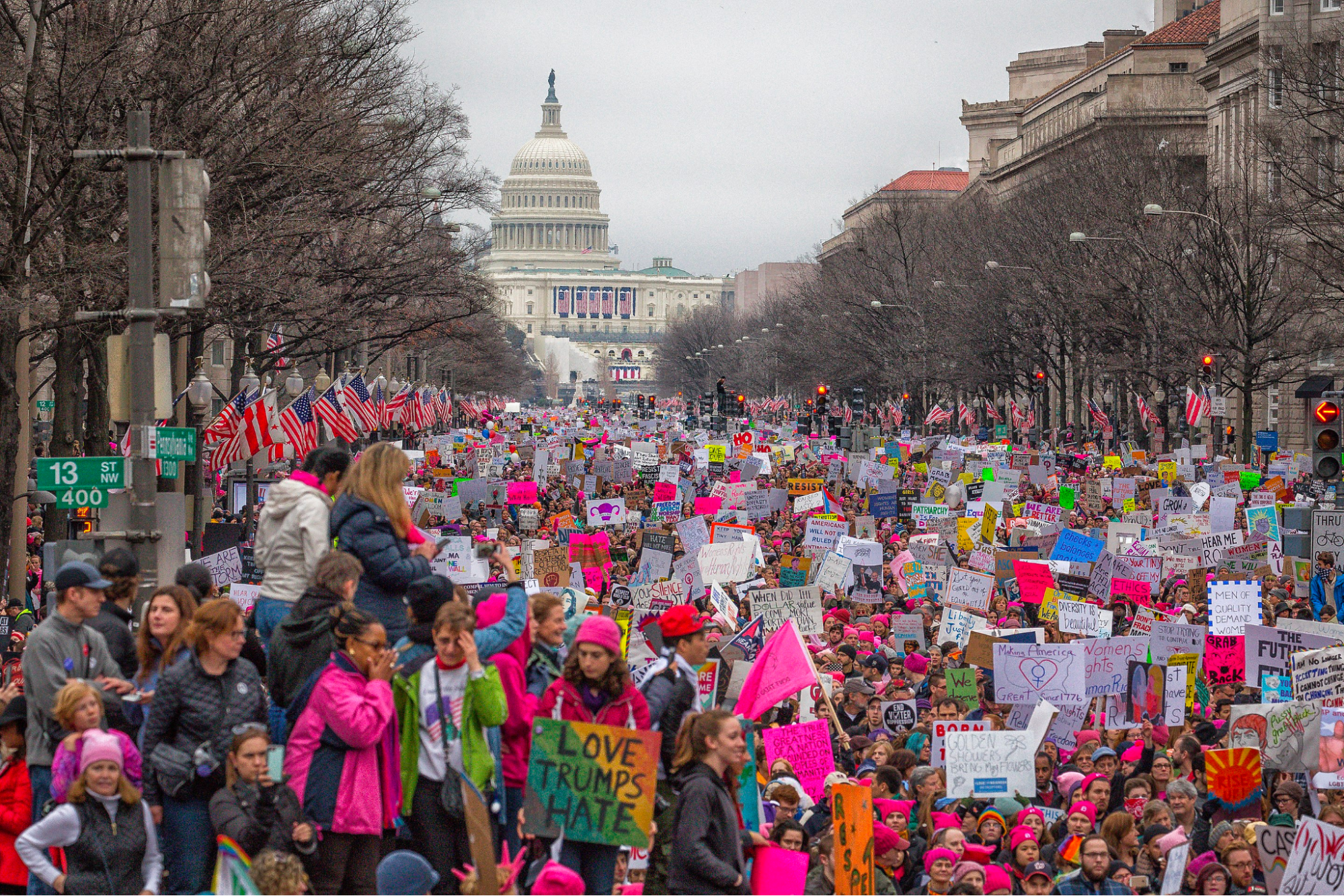 A very large group of people carrying sings and banners at the 2017 Women's March. May are wearing pink hats. The capitol Building is in the background.