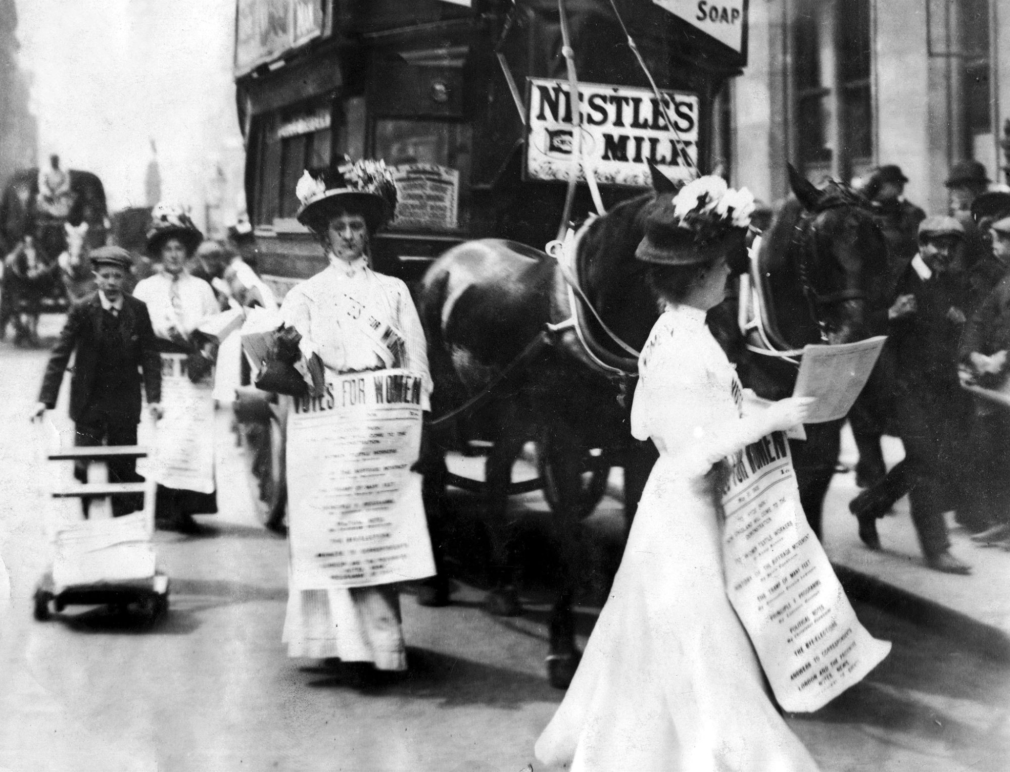 A vintage from the 1900's of women in with white dresses passing out fliers. They are wearing banners that say "Votes For Women"